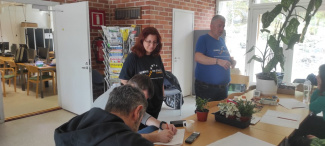 two persons standing in a room and talking, three persons sitting around a table and listening