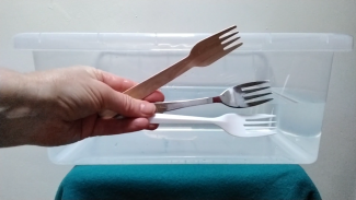 A hand is holding three different forks: one made of wood, one made of metal, one made of plastic. In the background, there is a water container.